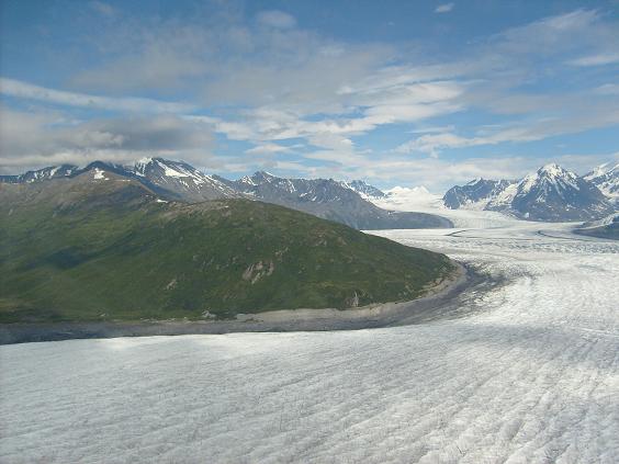 Glacier flowing between green mountains under a cloudy blue sky. Supermotors.net logo at bottom right