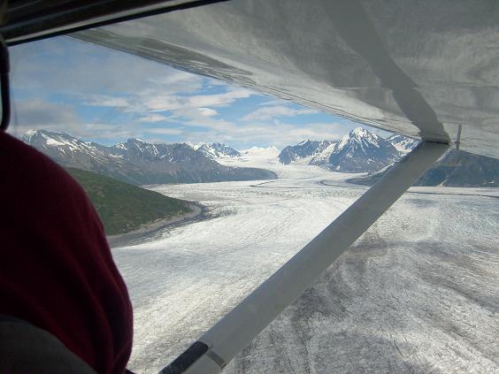 View of a vast glacier and snow-capped mountains from an airplane window, with a person's shoulder visible in the foreground