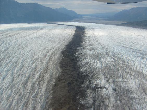 Aerial view of a vast glacier with a dark central moraine, mountains in the distance, and a portion of an aircraft wing at the top