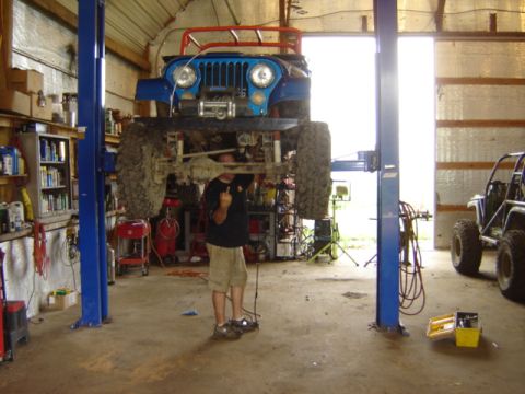 1992 Ford Bronco - Mechanic works on a lifted blue Jeep CJ in a cluttered garage bay.