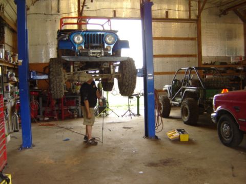 1992 Ford Bronco - A blue Jeep CJ-7 is suspended on a lift in a garage, with other vehicles visible in the background.