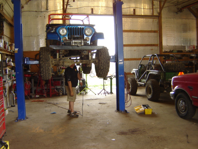 Blue Jeep CJ-7 on a lift, front view, with a red Ford F-Series truck and a green off-road vehicle in the background