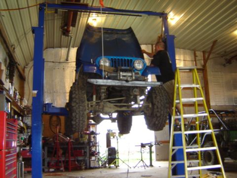 1992 Ford Bronco - A blue Jeep is suspended on a lift in a workshop, with its hood open and a mechanic attending to the engine.