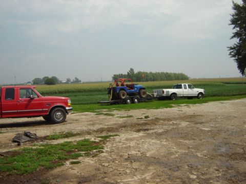 1992 Ford Bronco - A red pickup truck is parked on a dirt lot, towing a trailer with a blue off-road vehicle and a white pickup truck in the background.