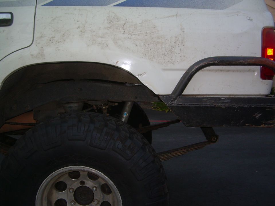 White SUV rear fender with large off-road tire, leaf spring suspension, and custom bumper. Scratches and dirt visible