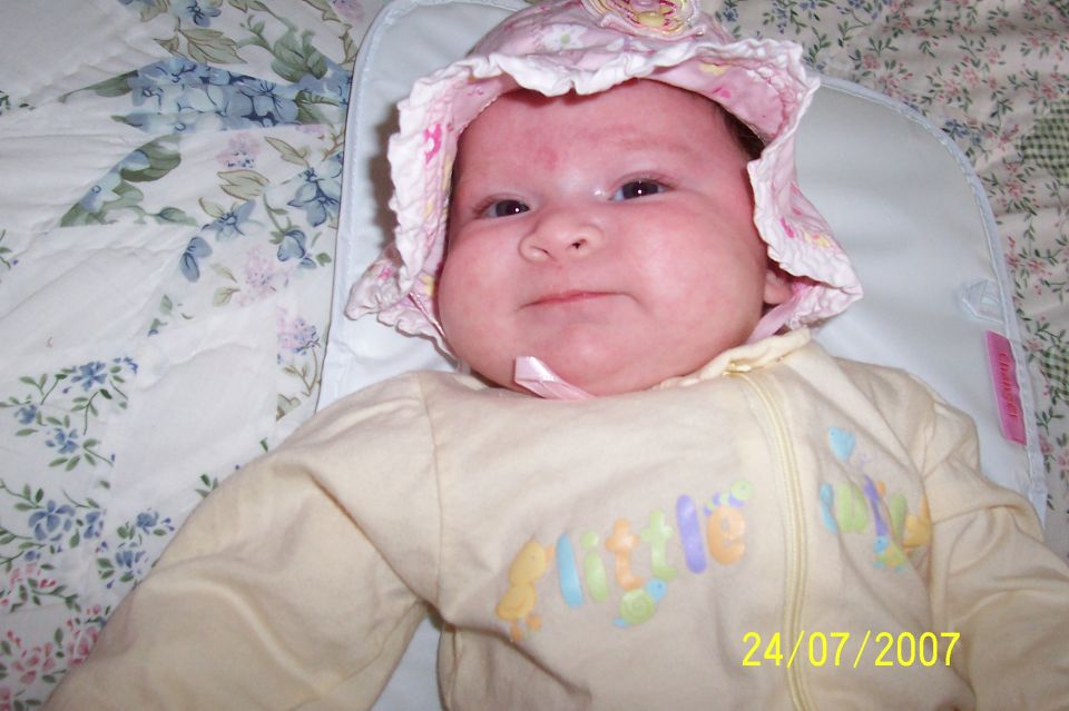 Baby wearing a pink bonnet and yellow onesie with colorful text, lying on a floral blanket. Date 24/07/2007 visible