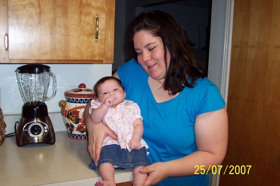 Woman in blue shirt holding a baby in a kitchen with a blender and decorative jar on the counter