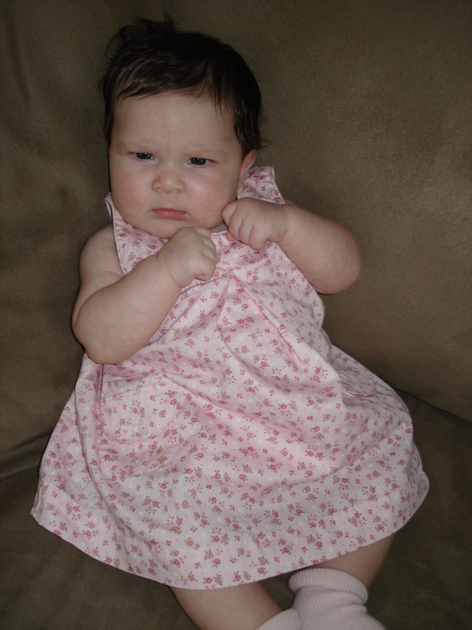 Baby in a pink floral dress and pink socks sits on a brown couch