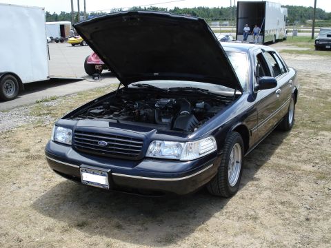 2000 Ford Crown Victoria - A dark blue 2000 Ford Crown Victoria is parked with its hood open, revealing the engine compartment. Other vehicles and trailers are visibl…