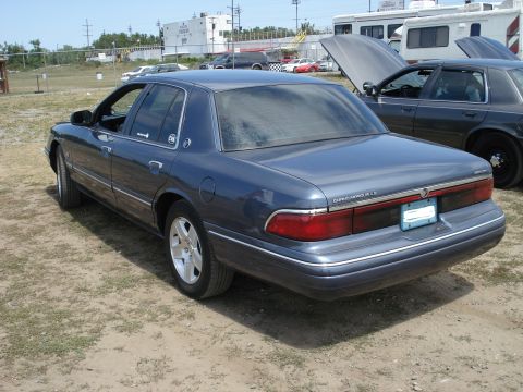 2000 Ford Crown Victoria - A dark blue Mercury Grand Marquis is parked on a dirt lot next to another vehicle with its hood open.