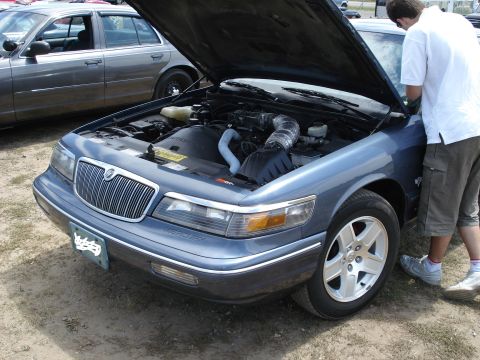 2000 Ford Crown Victoria - A blue Mercury Grand Marquis is shown with its hood open, revealing the engine compartment, while another vehicle sits in the background.