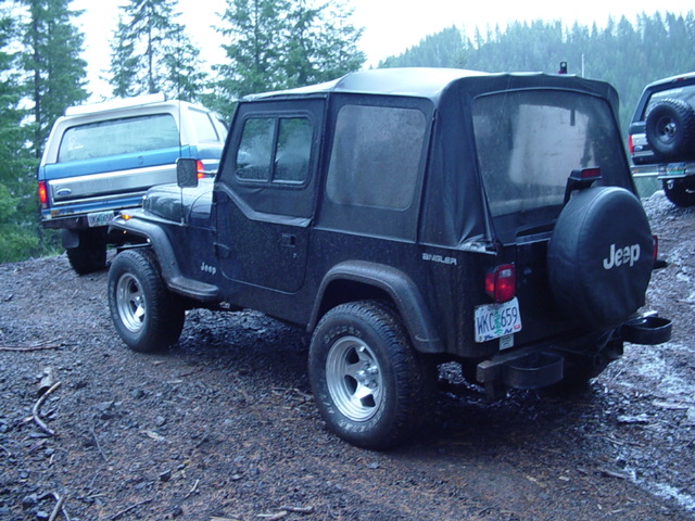 Black Jeep Wrangler with soft top and Jeep spare cover parked on a dirt road, trucks behind