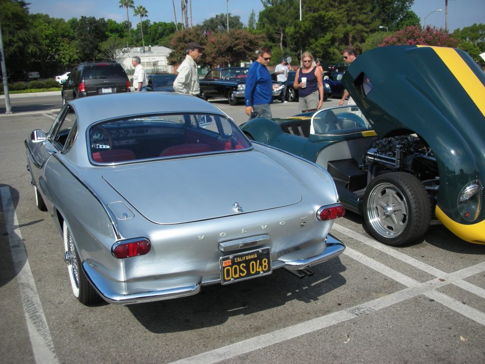 Silver Volvo P1800 coupe rear view, California license plate QQS 048. Green and yellow sports car with hood open next to it