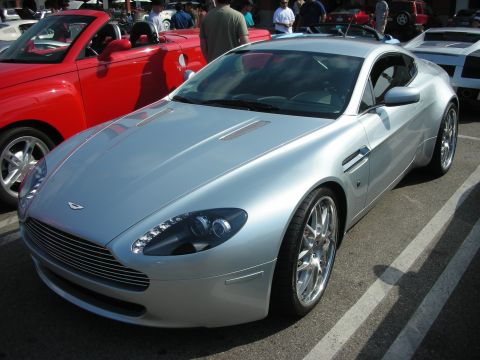 1998 Ford Crown Victoria - A silver Aston Martin coupe is parked at an outdoor car meet, with a red convertible beside it.