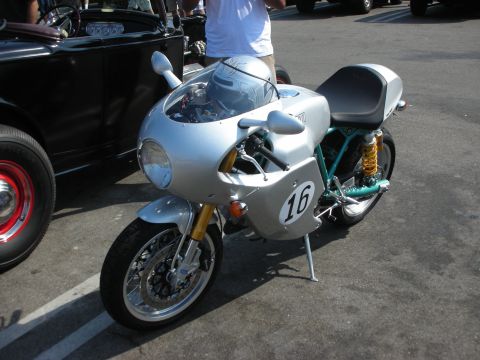 1998 Ford Crown Victoria - A silver cafe racer motorcycle, marked with the number 16, is parked next to a black classic car at an outdoor gathering.