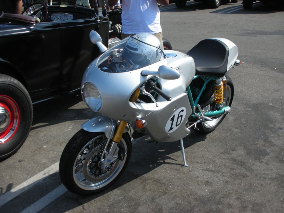 Silver cafe racer motorcycle with number 16 on side fairing, parked next to a black classic car