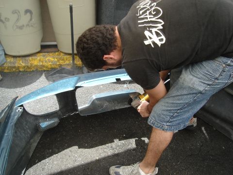 1995 Mazda MX-6 - Man modifies a car bumper with an angle grinder outdoors.
