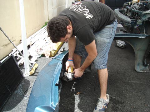 1995 Mazda MX-6 - Man working on a car bumper with an angle grinder, surrounded by vehicle parts.