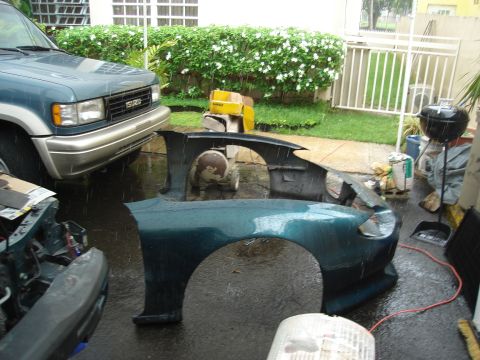 1995 Mazda MX-6 - Disassembled teal car body parts, including a fender and bumper, lie on wet asphalt beside an Isuzu SUV.
