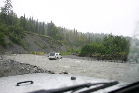 2003 Jeep TJ - A white SUV navigates a shallow river in a wooded, hilly area under an overcast sky.