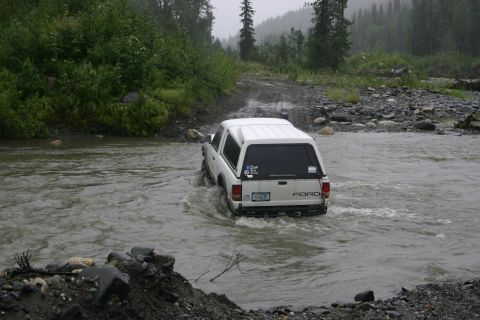 2003 Jeep TJ - A white Ford pickup truck with a camper shell navigates through a shallow river on a dirt road, with trees and rocky terrain in the backgro…