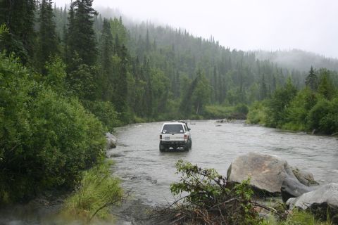 2003 Jeep TJ - SUV navigates a river crossing on a misty day, surrounded by dense forest.