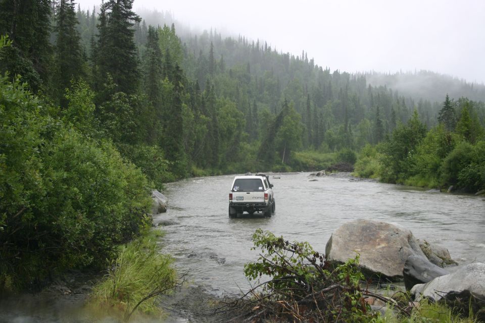 White SUV driving through a shallow river with forested hills in the background under a cloudy sky