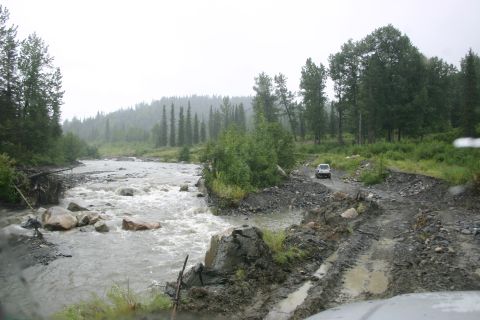 2003 Jeep TJ - A white SUV navigates a challenging muddy road alongside a turbulent river in a forested, hilly area.