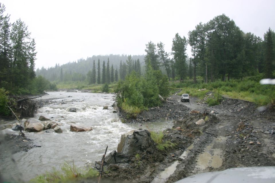 White SUV drives on a muddy, rutted road beside a fast-flowing river with trees and hills in the background