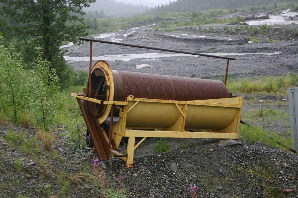 Rusty trommel screen with yellow frame on gravelly ground, trees and a riverbed in background