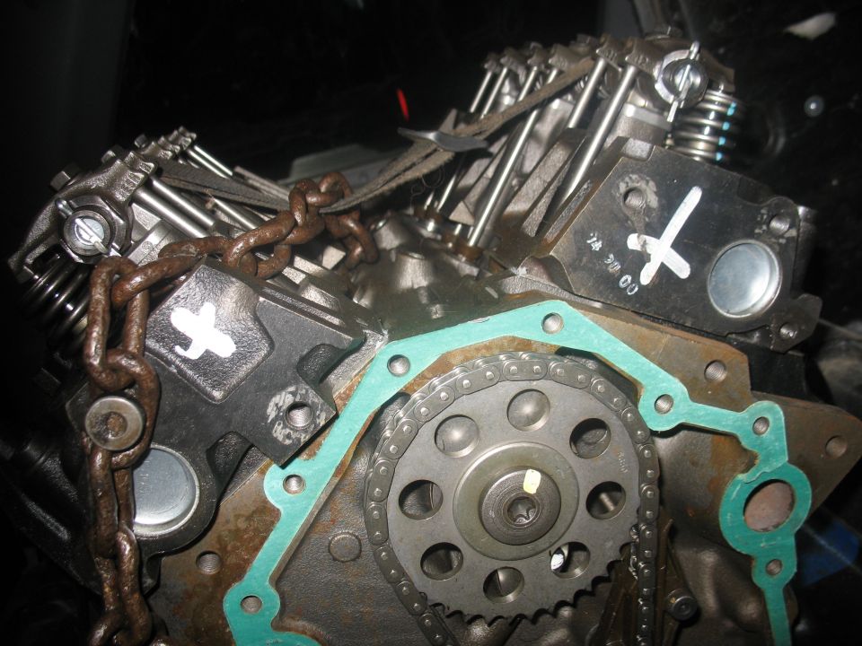 Engine block with timing chain, gasket, and chain tensioner visible. White 'X' marks on metal parts