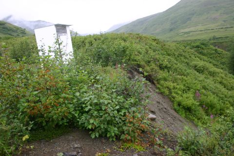 2003 Jeep TJ - A solitary outhouse stands in a lush, green valley under a cloudy sky.