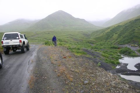 2003 Jeep TJ - A white Ford pickup truck with a camper shell drives on a wet dirt road through a valley with green hills and mountains.