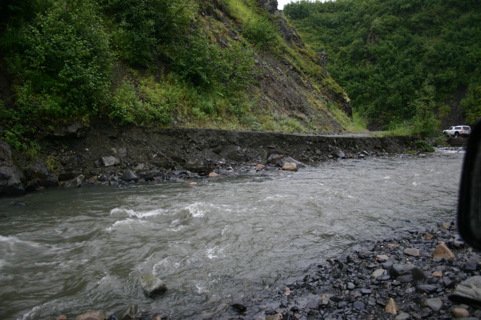 White SUV on a road damaged by a river, with trees on a hillside
