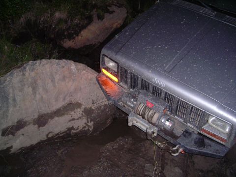 2003 Jeep TJ - A Jeep Cherokee XJ navigates a difficult off-road obstacle, getting stuck on a large rock in muddy terrain.