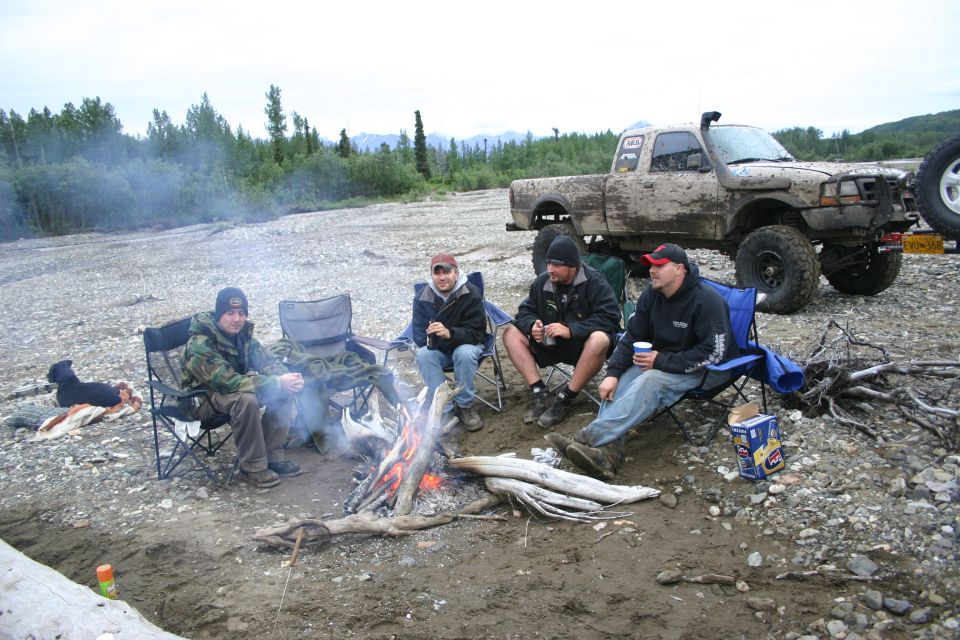 Mud-covered pickup truck with snorkel, parked on a rocky shore near a campfire with four people