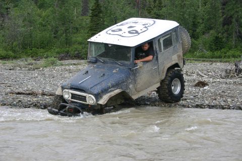 2003 Jeep TJ - A driver navigates a muddy Toyota Land Cruiser through a rocky riverbed surrounded by trees.
