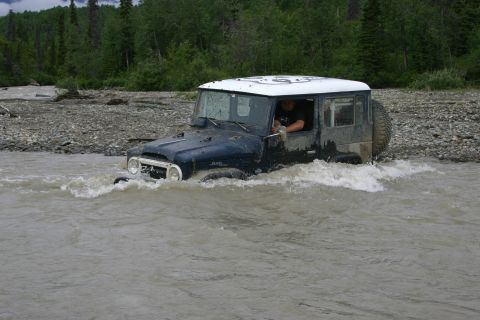 2003 Jeep TJ - A dark blue 2003 Jeep Wrangler TJ navigates through a shallow river with trees and rocky terrain in the background.