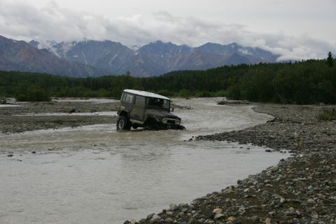 2003 Jeep TJ - An off-road vehicle navigates a rocky riverbed under a cloudy sky with distant mountains.