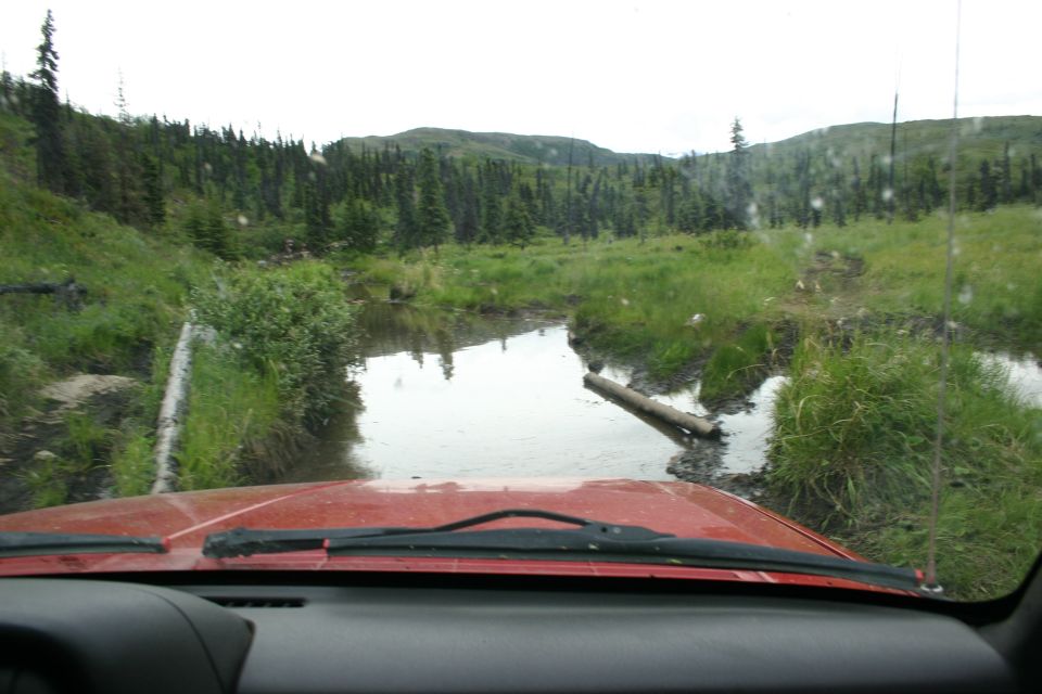 Red vehicle hood and windshield viewed from interior, driving through a muddy, wet terrain with trees and hills in background