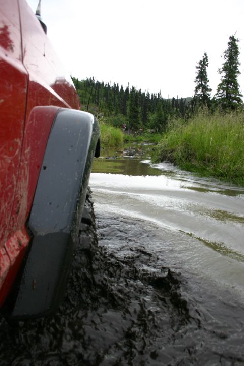 2003 Jeep TJ - A red 2003 Jeep TJ navigates a muddy trail, kicking up water as it drives through the obstacle.