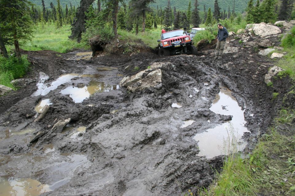 Red Jeep Wrangler navigating a very muddy off-road trail with deep ruts and puddles. Another vehicle is visible behind it