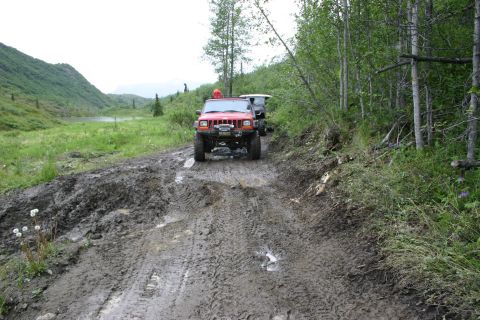 2003 Jeep TJ - Two SUVs navigate a challenging muddy trail with dense forest on the right and a lake on the left.