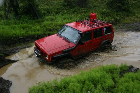 2003 Jeep TJ - A red Jeep Cherokee XJ navigates a challenging muddy water crossing, equipped for off-road adventure.