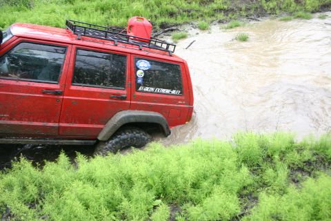 2003 Jeep TJ - A red Jeep Cherokee XJ navigates through a muddy water obstacle, equipped with a roof rack and a jerry can.