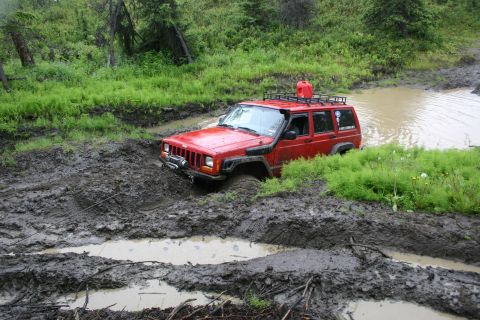 2003 Jeep TJ - A red Jeep Cherokee navigates a challenging muddy off-road course, with its tires deep in the muck.