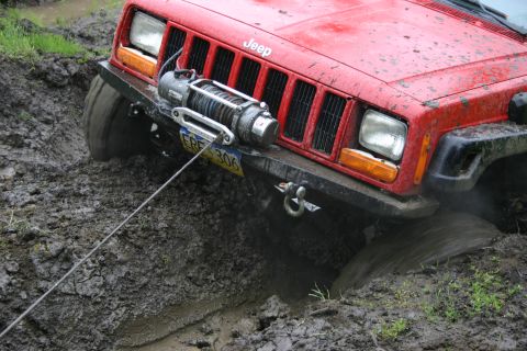 2003 Jeep TJ - A red Jeep Cherokee XJ is stuck in thick mud, its front tire submerged and a winch cable extending from the bumper.