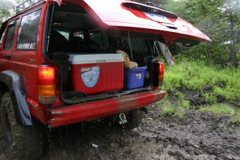 2003 Jeep TJ - The rear of a red Jeep Cherokee XJ is shown with its tailgate open, loaded with a cooler and a blue bin, in a muddy, wooded area.