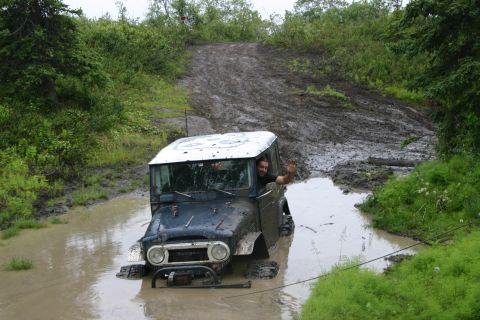 2003 Jeep TJ - A driver waves from a dark blue off-road vehicle that is stuck in a muddy puddle on a trail.