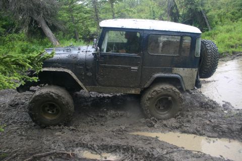 2003 Jeep TJ - A heavily mud-splattered off-road vehicle navigates a difficult muddy terrain surrounded by trees.